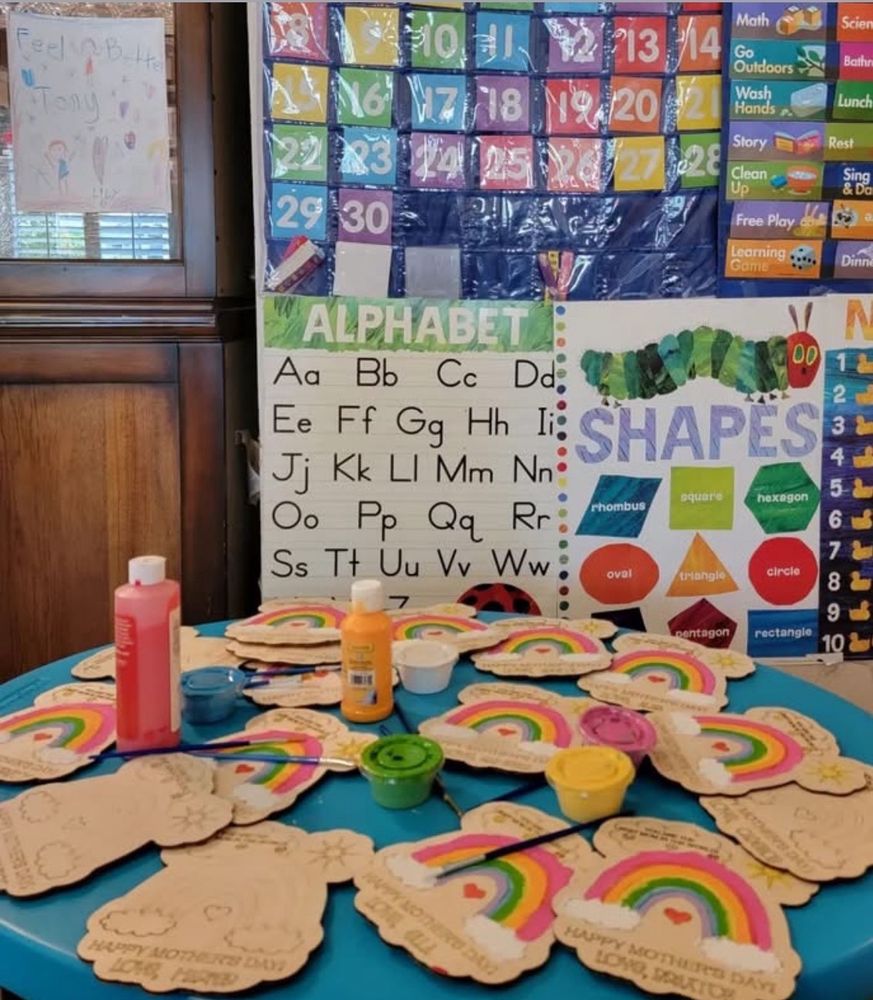 The learning corner with painted rainbow craft cutouts on a teal table, an alphabet chart, a calendar and a daily schedule on the wall